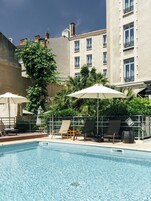 Una piscina con sillas de descanso y sombrillas, rodeada de vegetación y edificios.