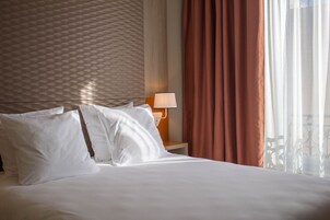 A hotel room with a bed, pillows, a nightstand with a lamp, and a window with curtains.