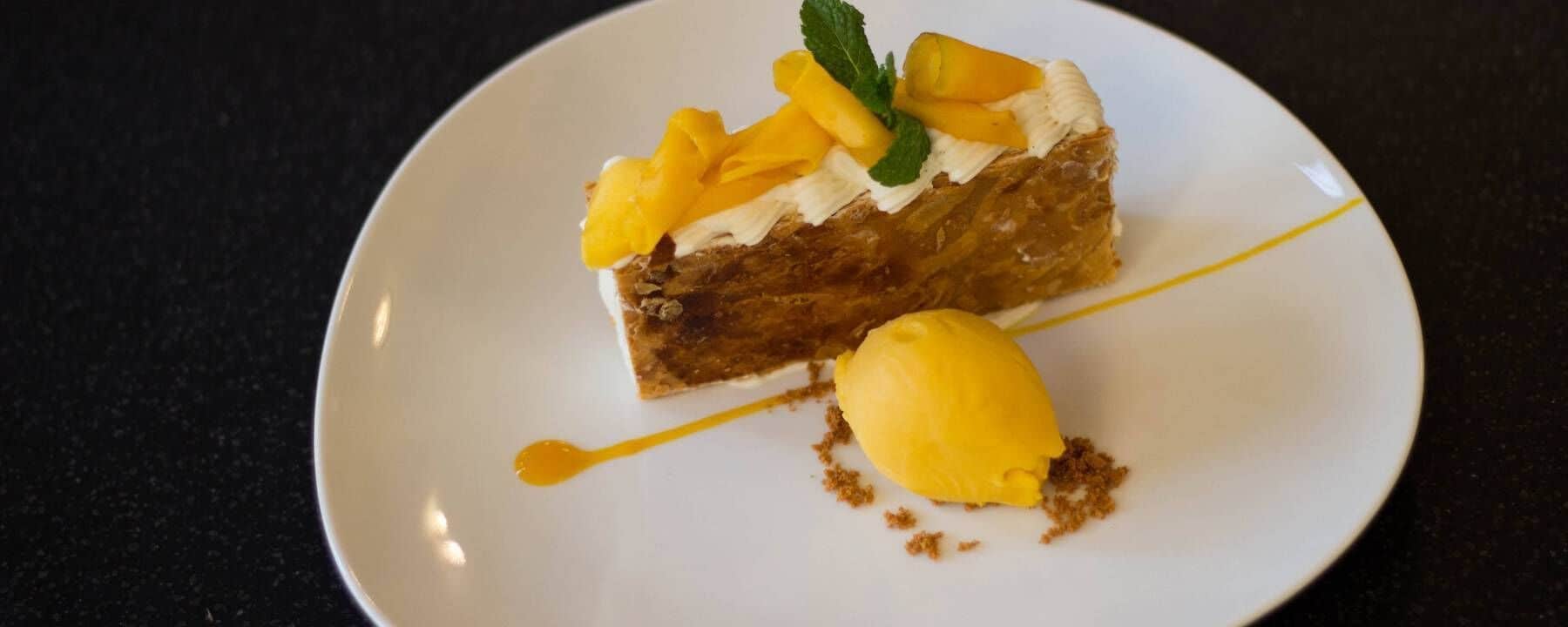 A slice of cake with mango topping and a scoop of yellow ice cream on a white plate.