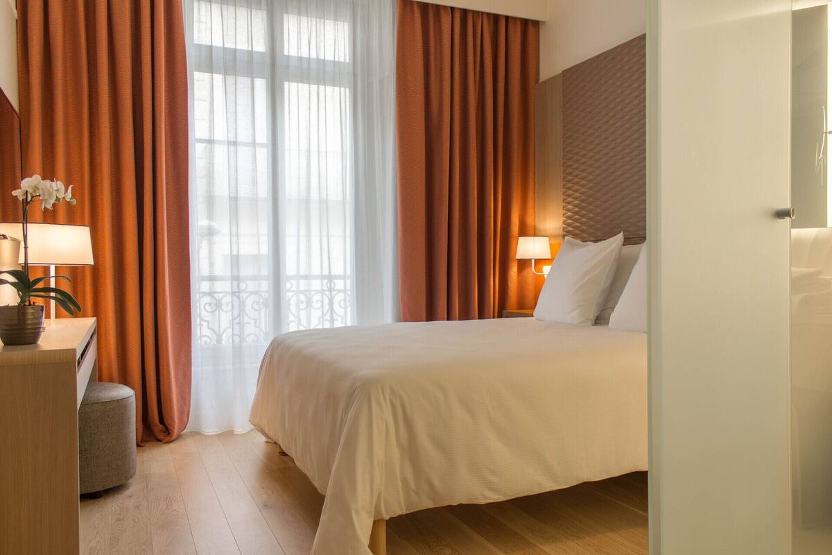 Een hotelkamer met een bed, twee bedlampen, een plant in pot en een raam met gordijnen.