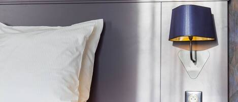 Una cama bien hecha con una colcha blanca y almohadas, una mesita de noche fijada a la pared con una lámpara, y un papel tapiz estampado.