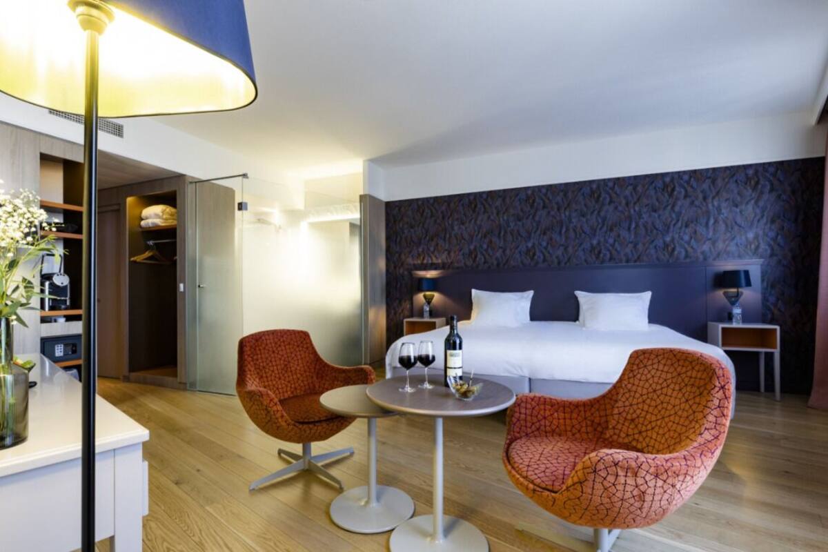 Een hotelkamer met een bed, twee fauteuils, een klein rond tafeltje met wijnglazen, een lamp en een kledingkast.