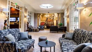 A spacious lobby with a grand staircase, comfortable seating, and a television displaying a news program. - Hôtel Oceania Le Métropole Montpellier (Montpellier)