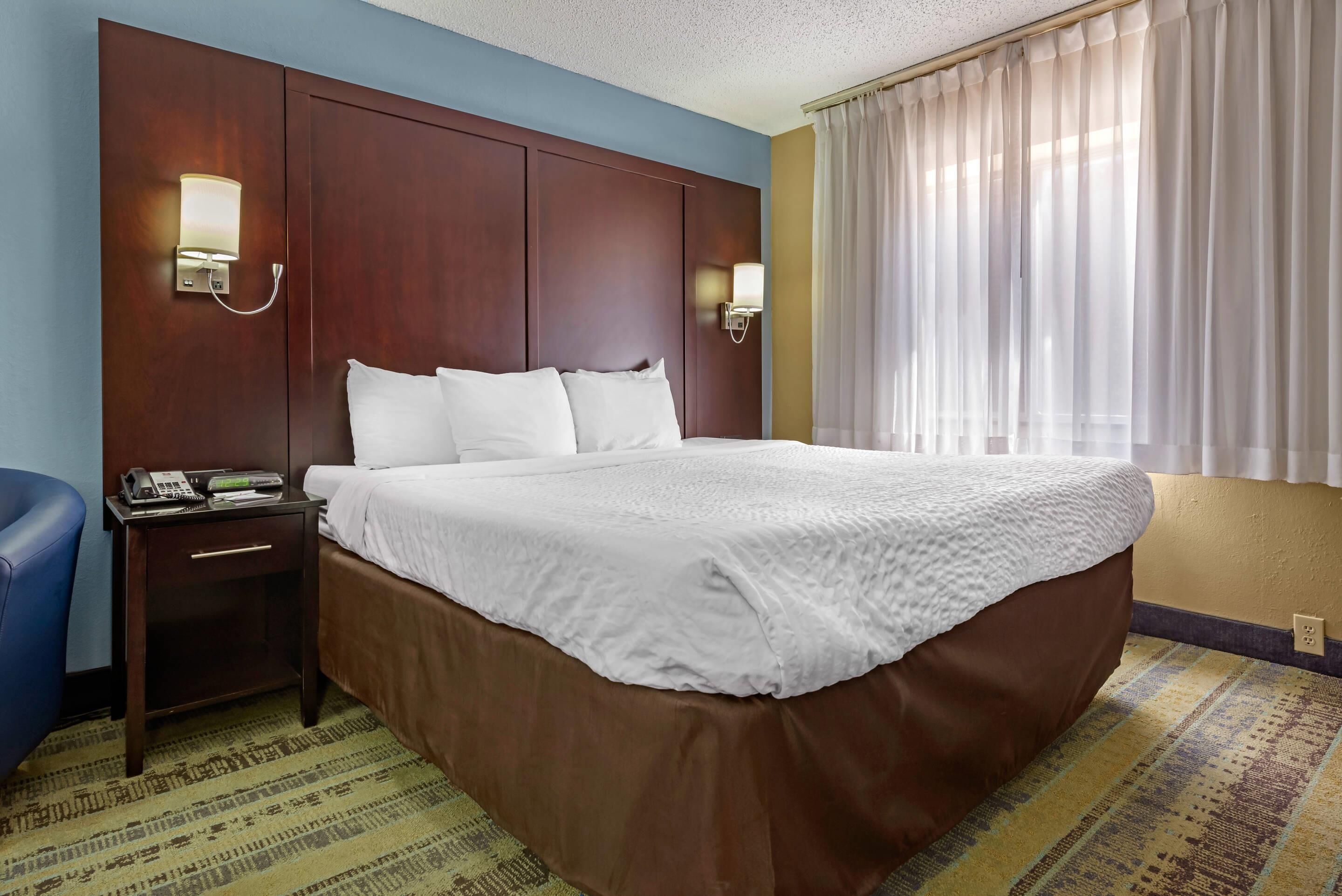 A hotel room with a large bed, a nightstand with a phone and a lamp, a blue chair, and a window with curtains.
