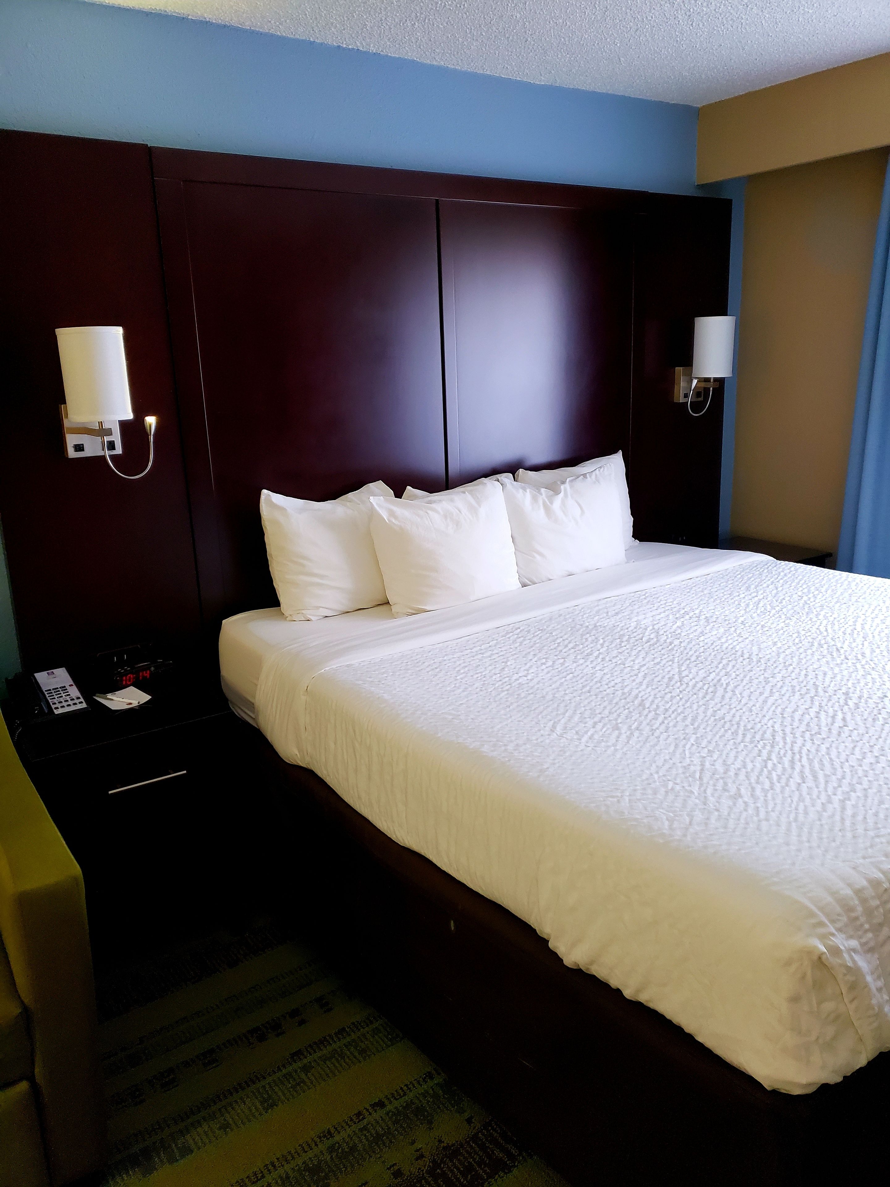 A hotel room with a large bed, dark headboard, bedside lamps, a nightstand with a phone and a clock, and a patterned carpet.