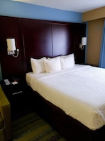 A hotel room with a large bed, dark headboard, bedside lamps, a nightstand with a phone and a clock, and a patterned carpet.