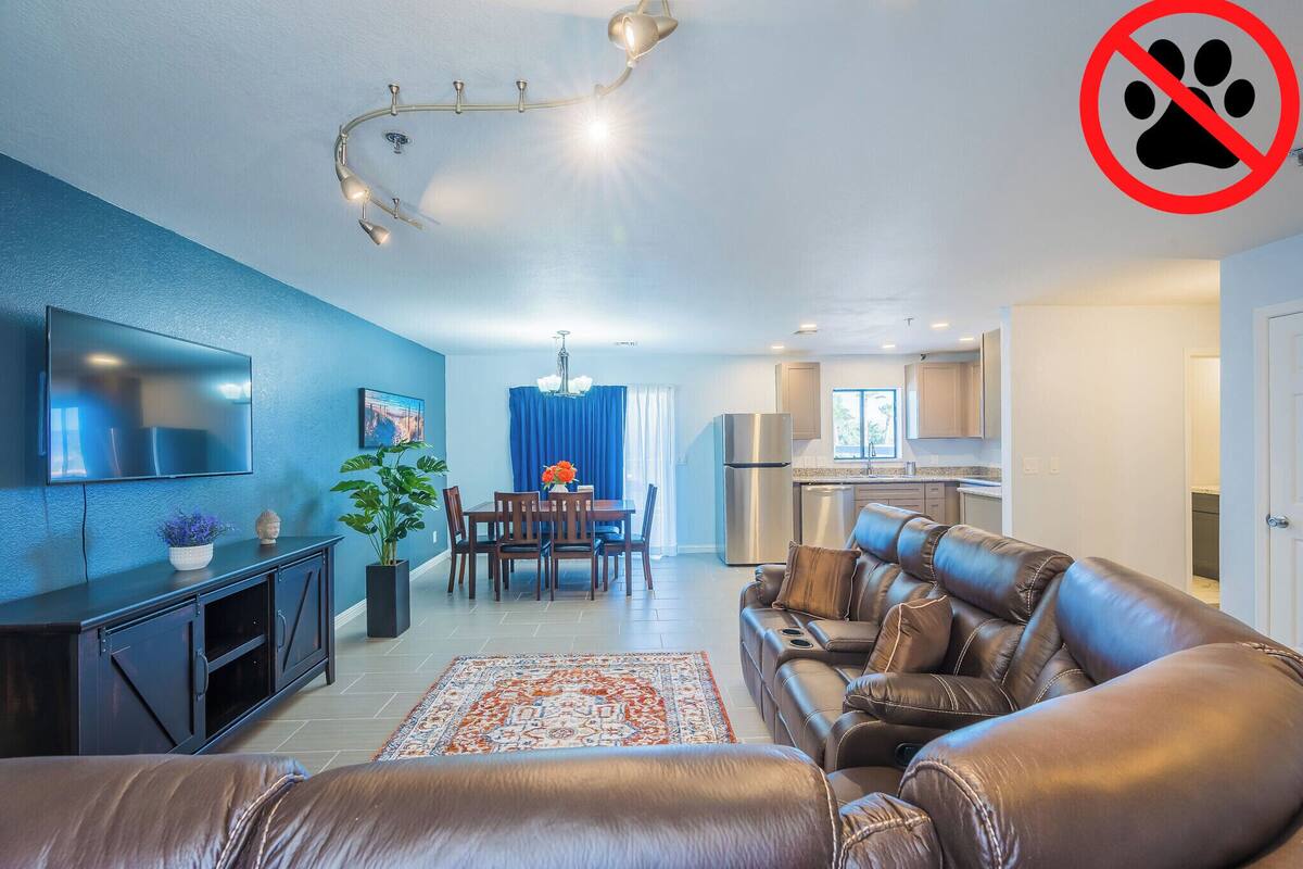 Una sala moderna con un sofá de cuero, un televisor de pantalla plana, una zona de comedor con mantel azul y una cocina al fondo.