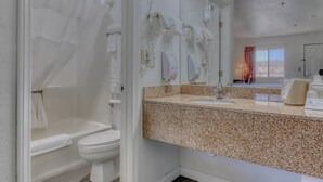 A bathroom with a white toilet, a shower with a transparent curtain, a granite countertop, and a mirror with lights.