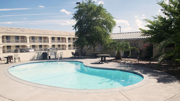 A swimming pool with a curved edge, surrounded by a concrete deck. There are outdoor tables and chairs, and a building with a tiled roof in the background.