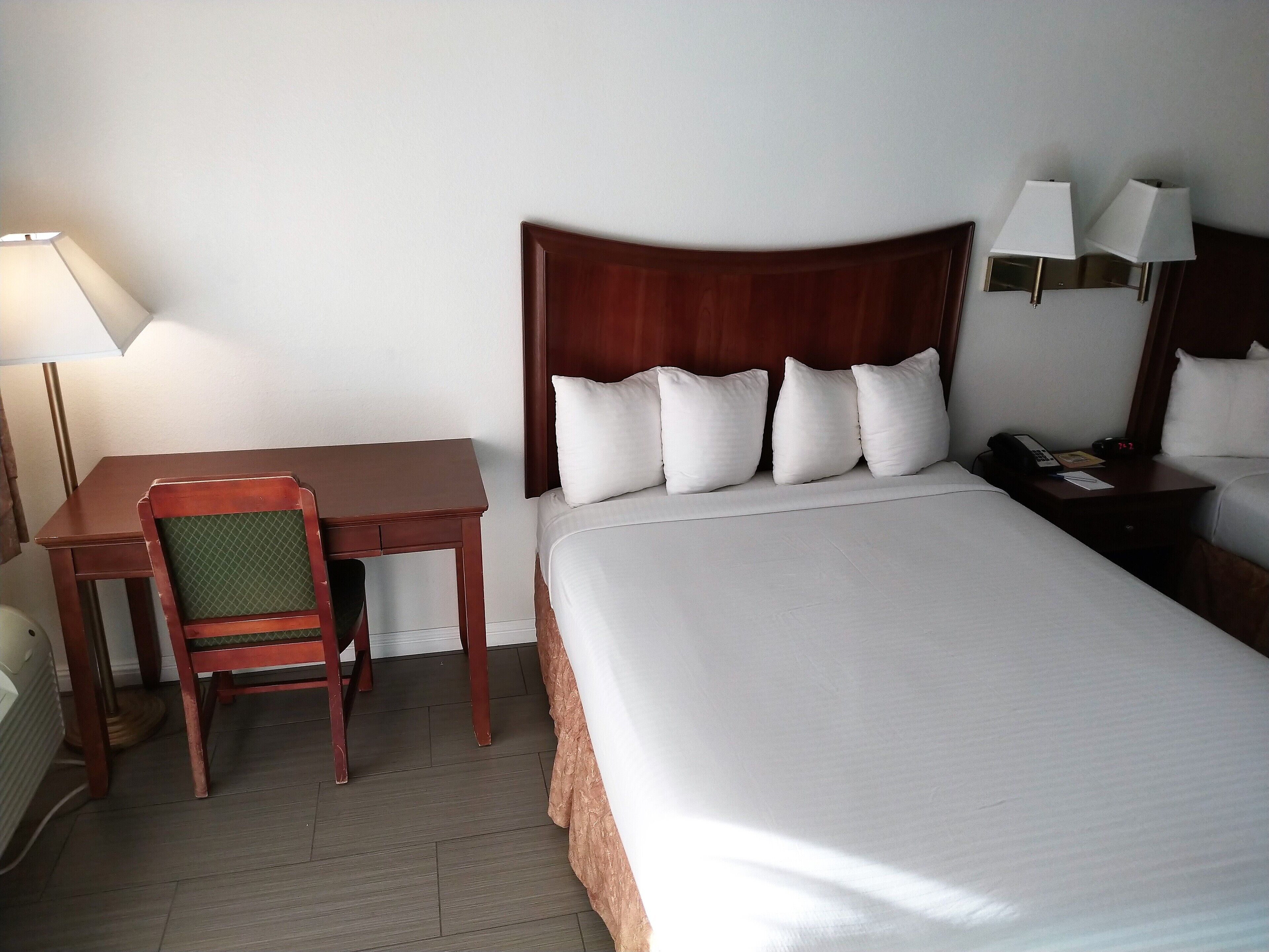 Una habitación de hotel con cama, escritorio, silla, lámpara y mesita de noche con teléfono y reloj.