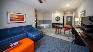 Habitación de hotel con sofá azul, otomana naranja, escritorio con computadora, televisor, ventilador de techo, un cuadro, área de cocina con lavamanos y una bañera.