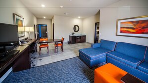 Una habitación de hotel moderna con televisor, escritorio, silla de oficina, mesa de comedor y sofá azul con detalles naranja.