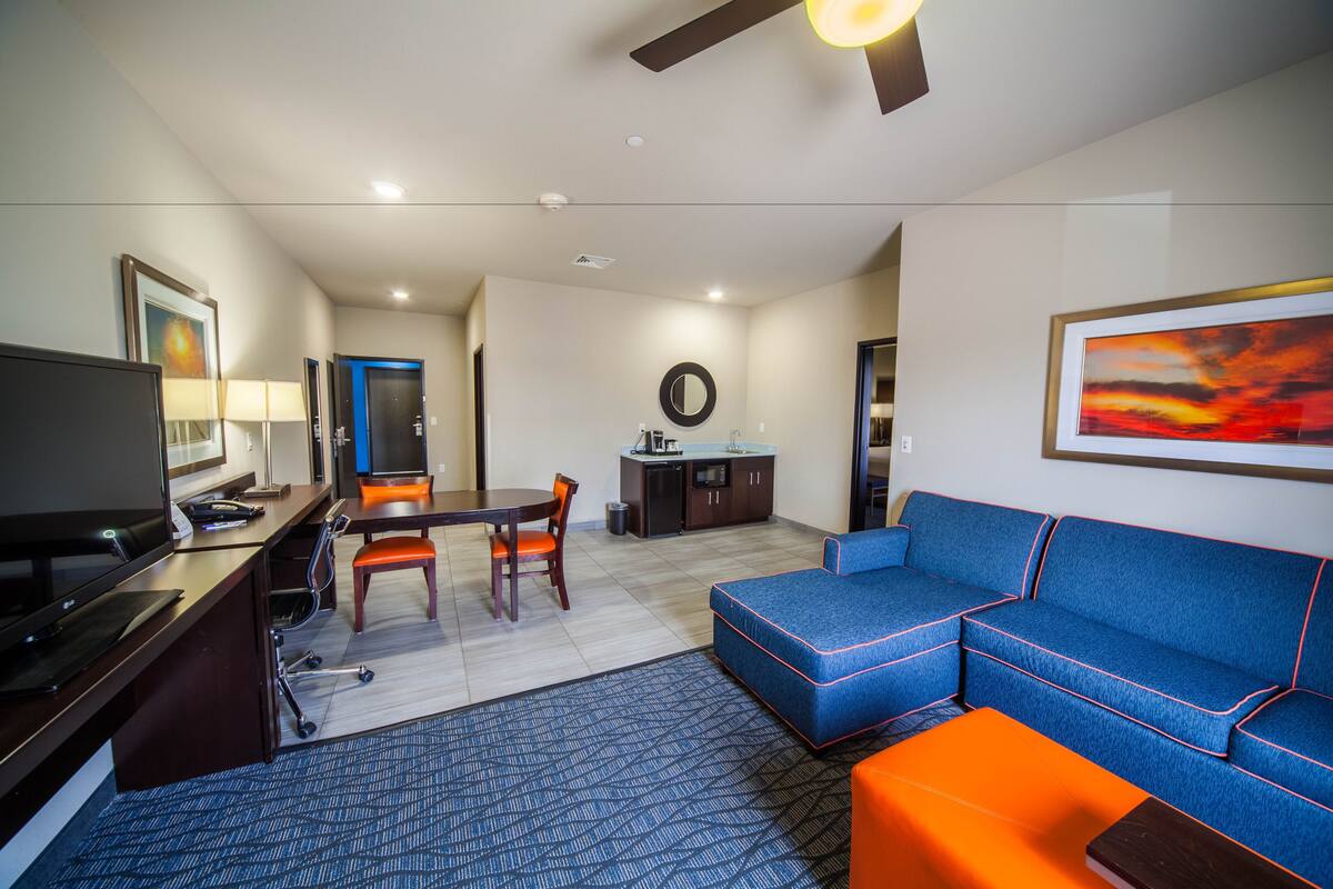 Ein modernes Hotelzimmer mit Fernseher, Schreibtisch, Bürostuhl, Esstisch und einem blauen Sofa mit orangefarbenen Akzenten.