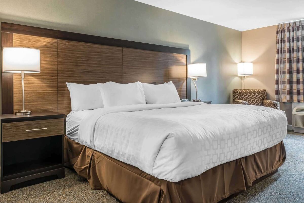 Quarto de hotel com uma cama grande, duas mesas de cabeceira com abajures, uma cadeira e ar-condicionado fixado na parede.
