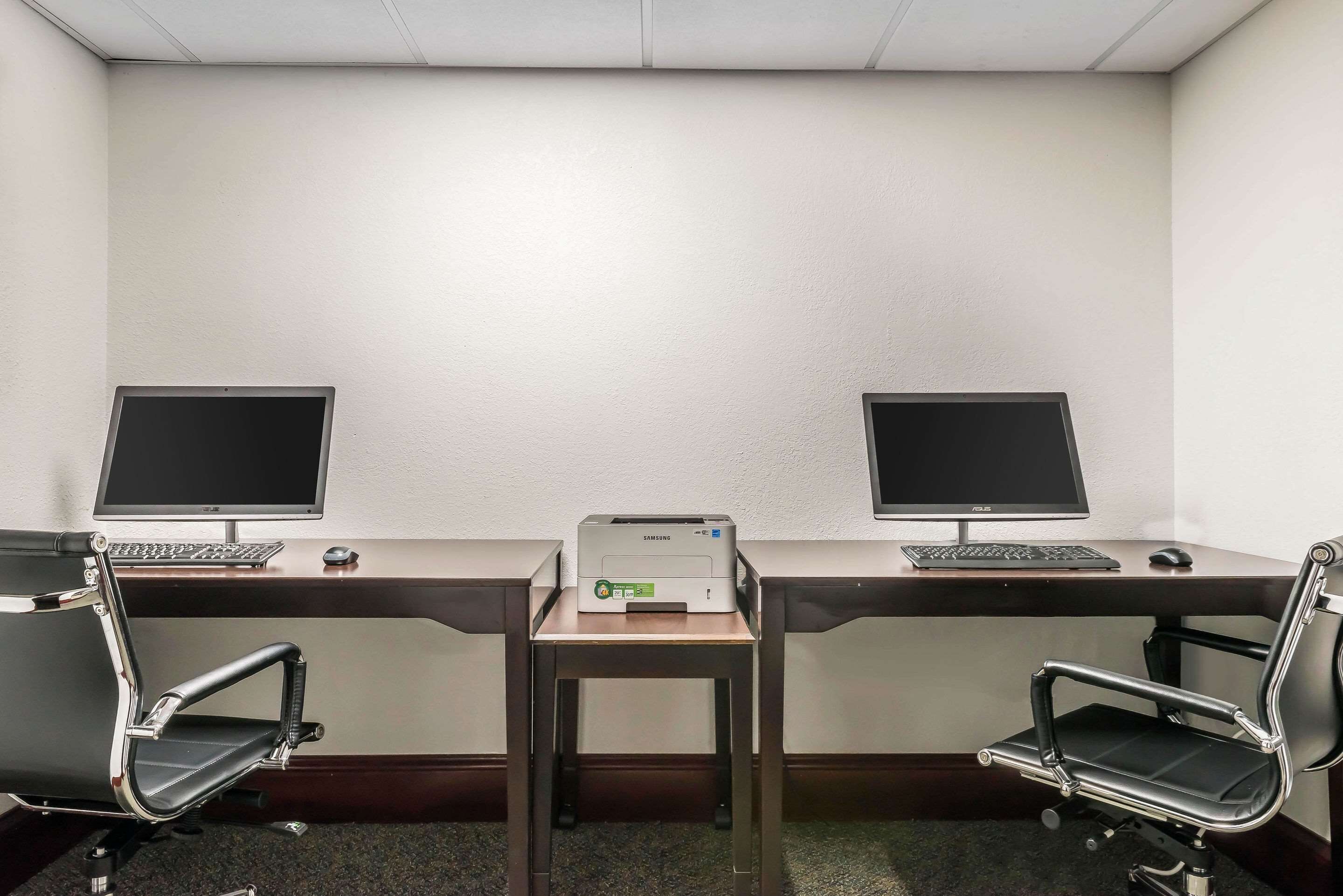 Et lille kontor med to skriveborde, hvert med computerskærm, tastatur og mus. To kontorstole, en ved hvert skrivebord. En printer står på gulvet mellem bordene.