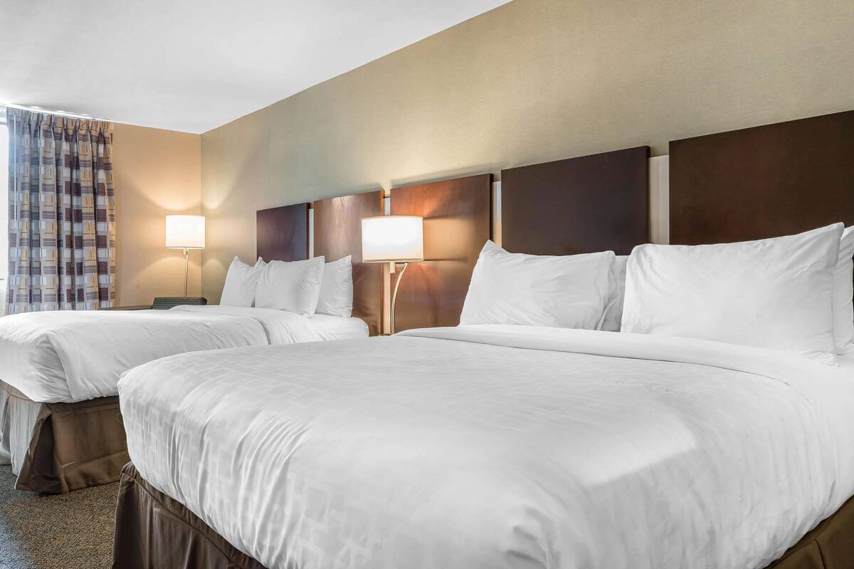 Quarto de hotel com duas camas, uma mesa de cabeceira, uma luminária e uma janela com cortinas.