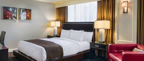 A hotel room with a large bed, two bedside tables with lamps, a red leather sofa, and a large window with curtains.
