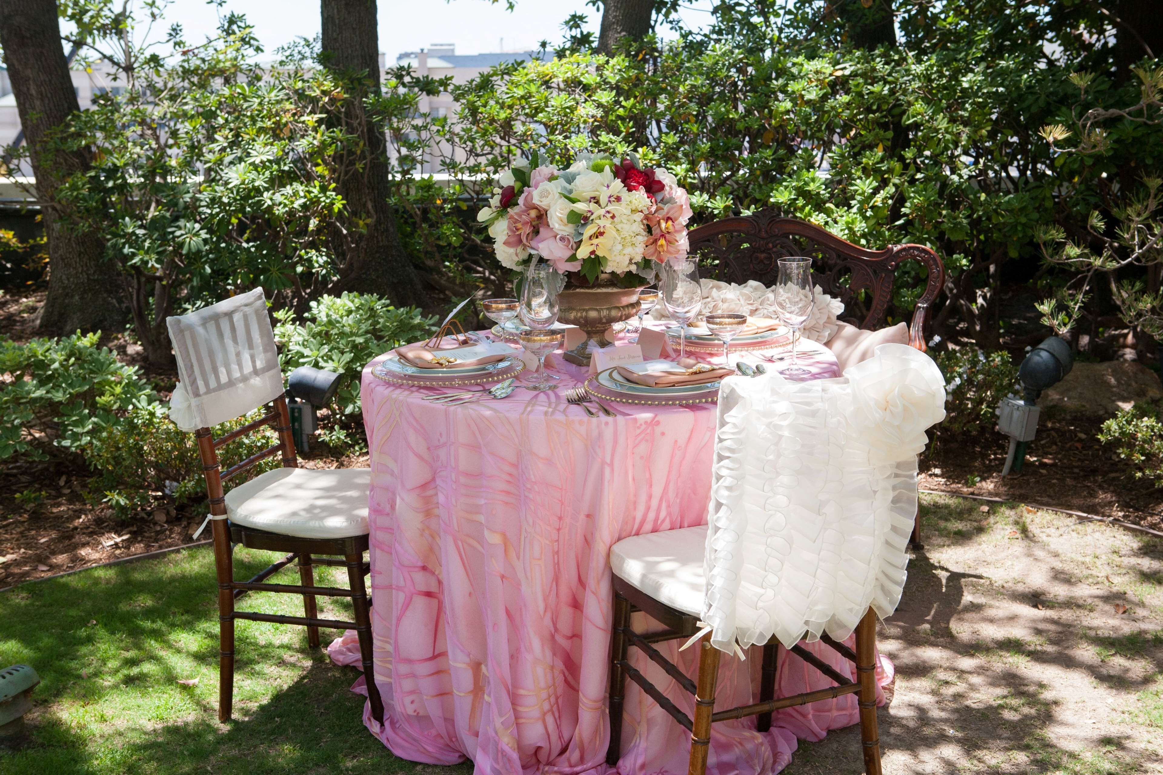 An outdoor dining table set with pink tablecloth, white chairs, and a floral centerpiece.