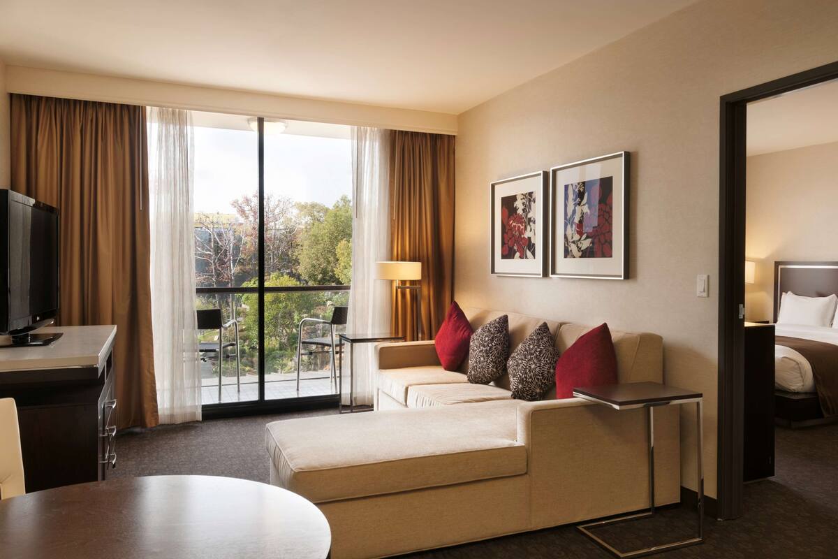Camera d'albergo con divano, televisione, scrivania, tavolo da pranzo e letto. Due quadri appesi al muro e un balcone con vista sugli alberi.