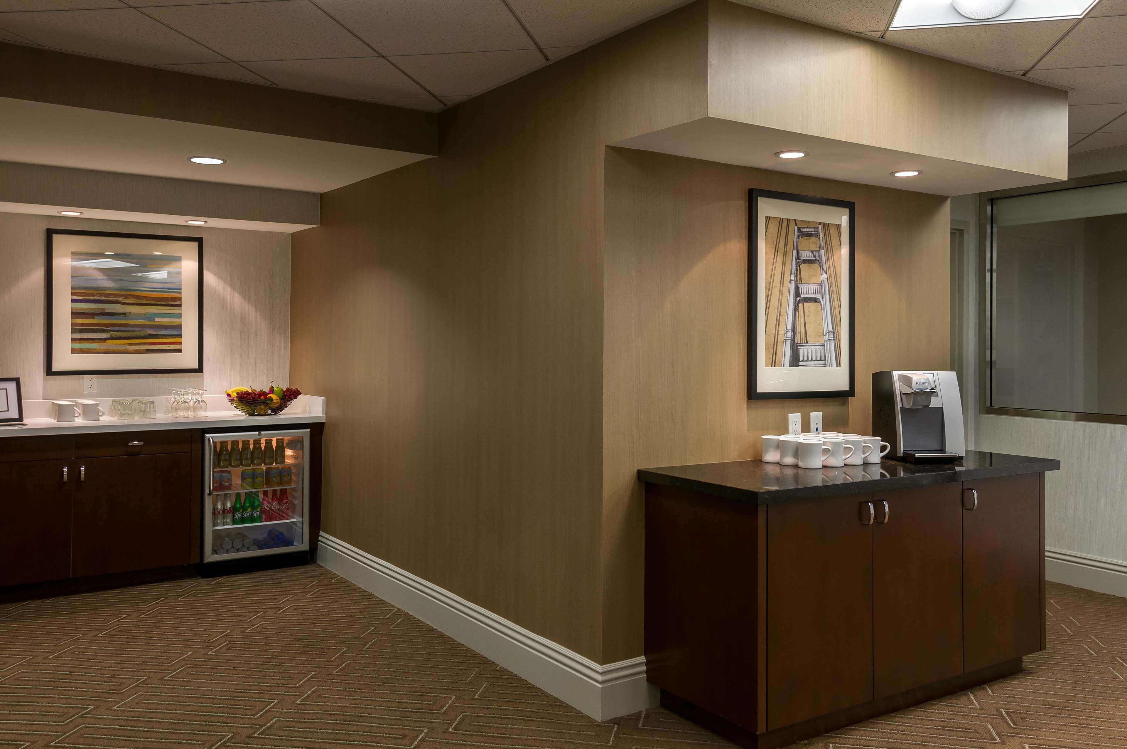 A hotel reception area with a refrigerated beverage dispenser, a coffee machine, and framed artwork on the walls.