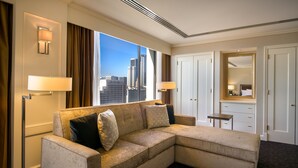 A hotel room with a beige sectional sofa, two wall-mounted lamps, a small bedside table, a mirror, and a view of the city skyline through large windows.