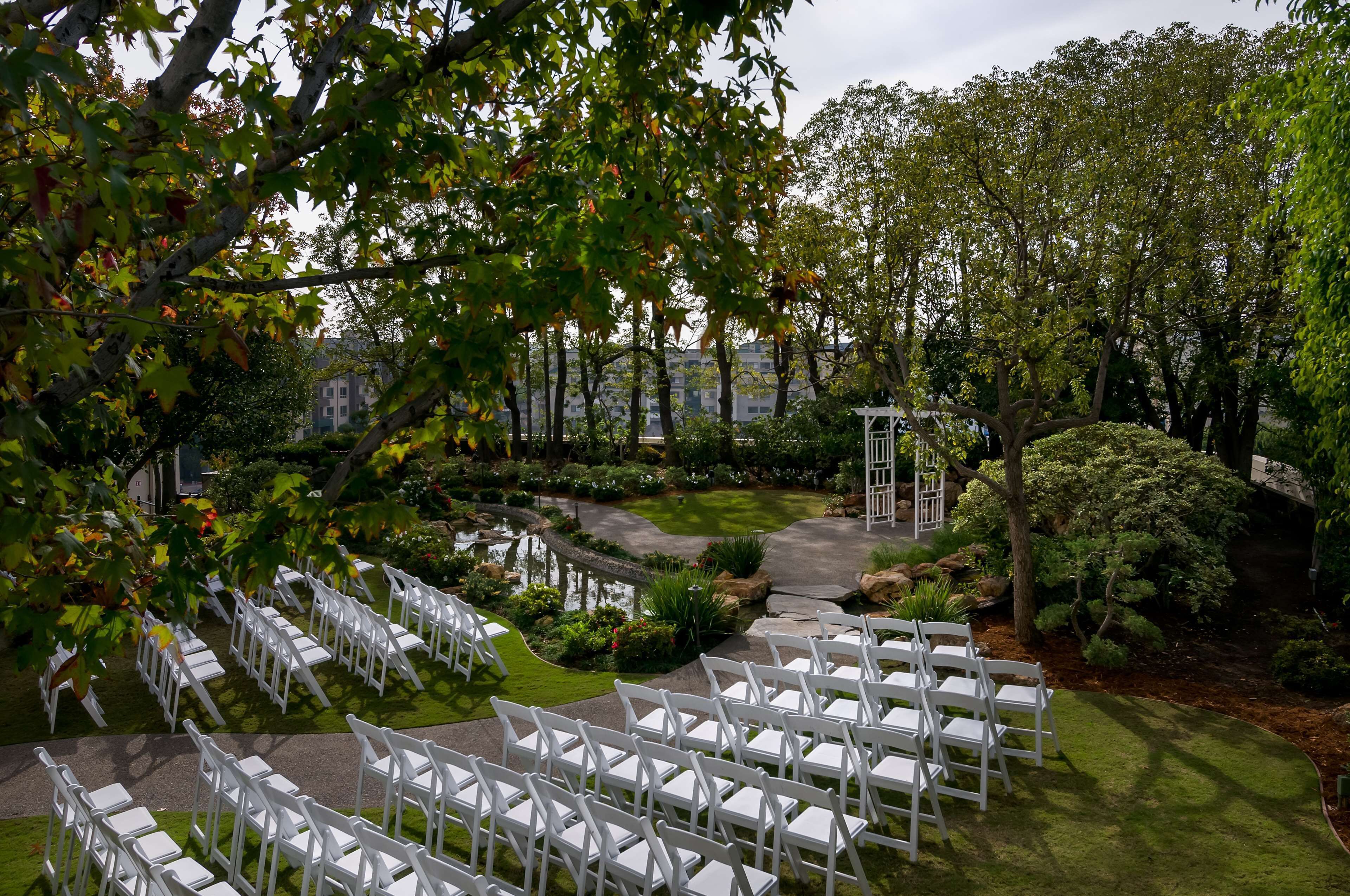 Un espacio al aire libre con jardín, con sillas blancas dispuestas para un evento, rodeado de vegetación exuberante y un pequeño estanque.