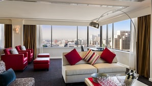 A modern hotel room with a sofa set, a dining table with wine glasses, and a city view.
