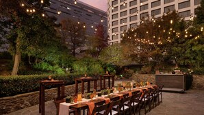 An outdoor dining area with tables set for a meal, surrounded by stone walls and greenery, with string lights overhead.