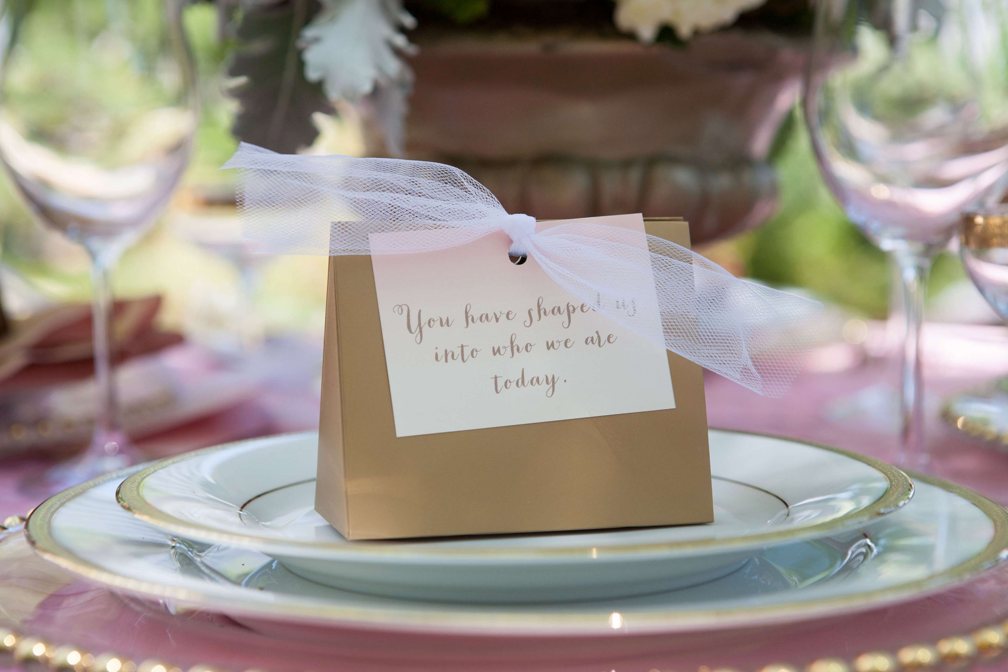 A small brown gift box with a white ribbon and a card on top, placed on a white plate with a gold rim, set on a table with a pink tablecloth.