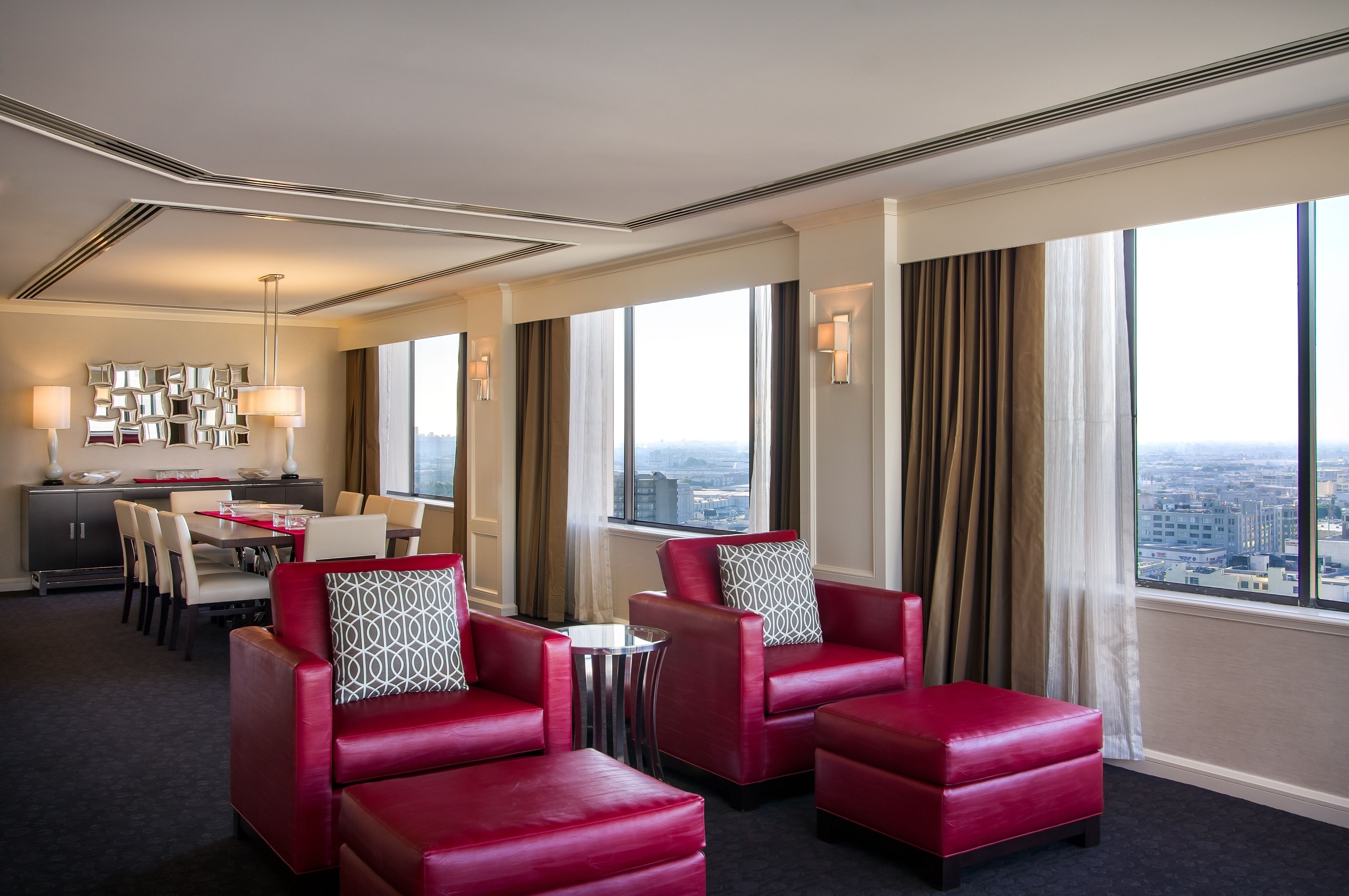 A hotel room with a city view, red leather chairs, a dining area, and a modern ceiling design.