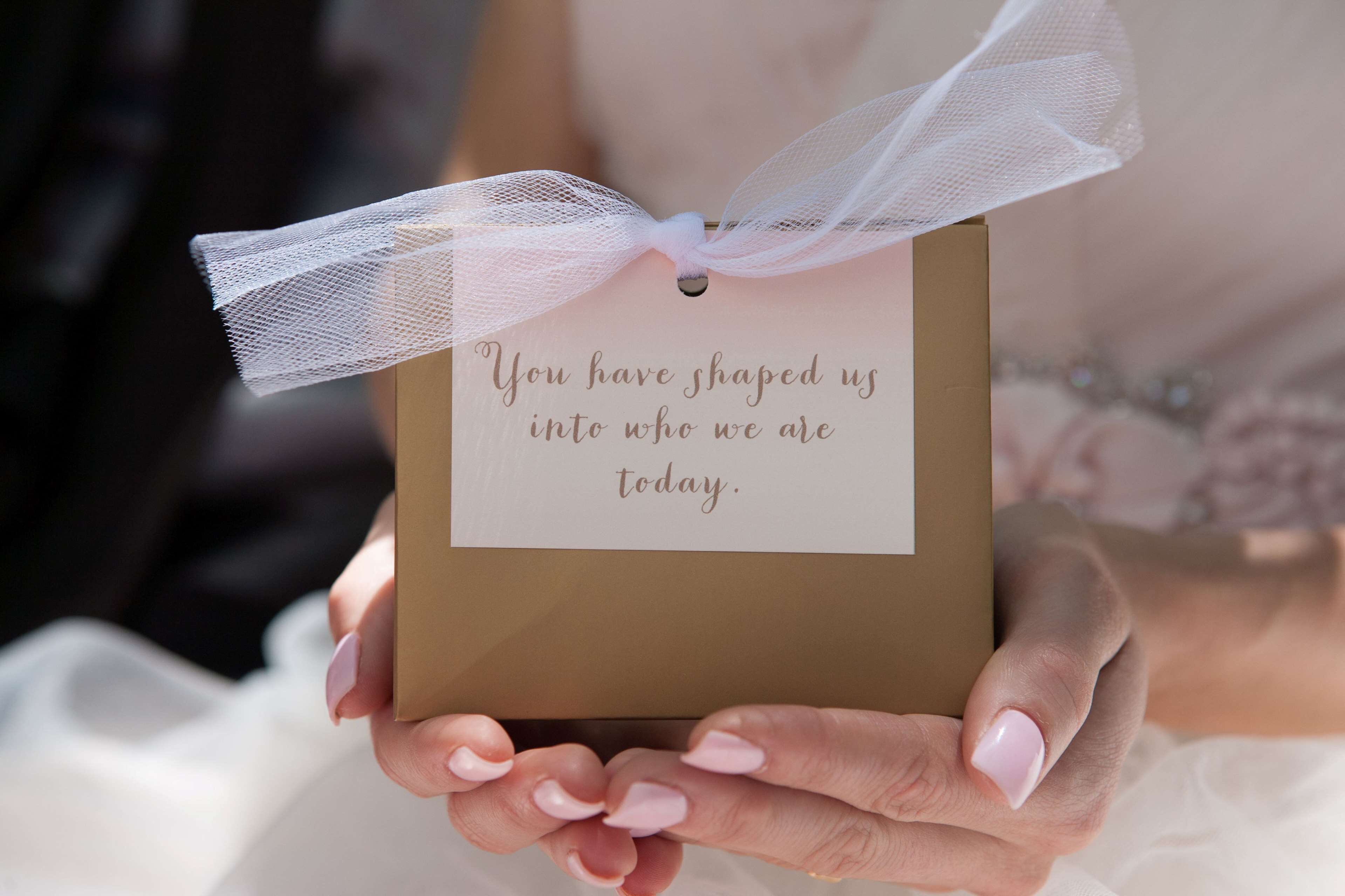 A person holding a small gift box with a ribbon and a card that reads "You have shaped us into who we are today."