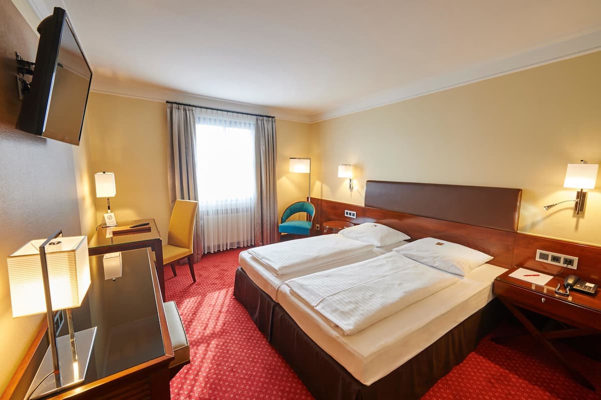 A hotel room with two beds, a desk with a lamp, a chair, and a TV mounted on the wall.