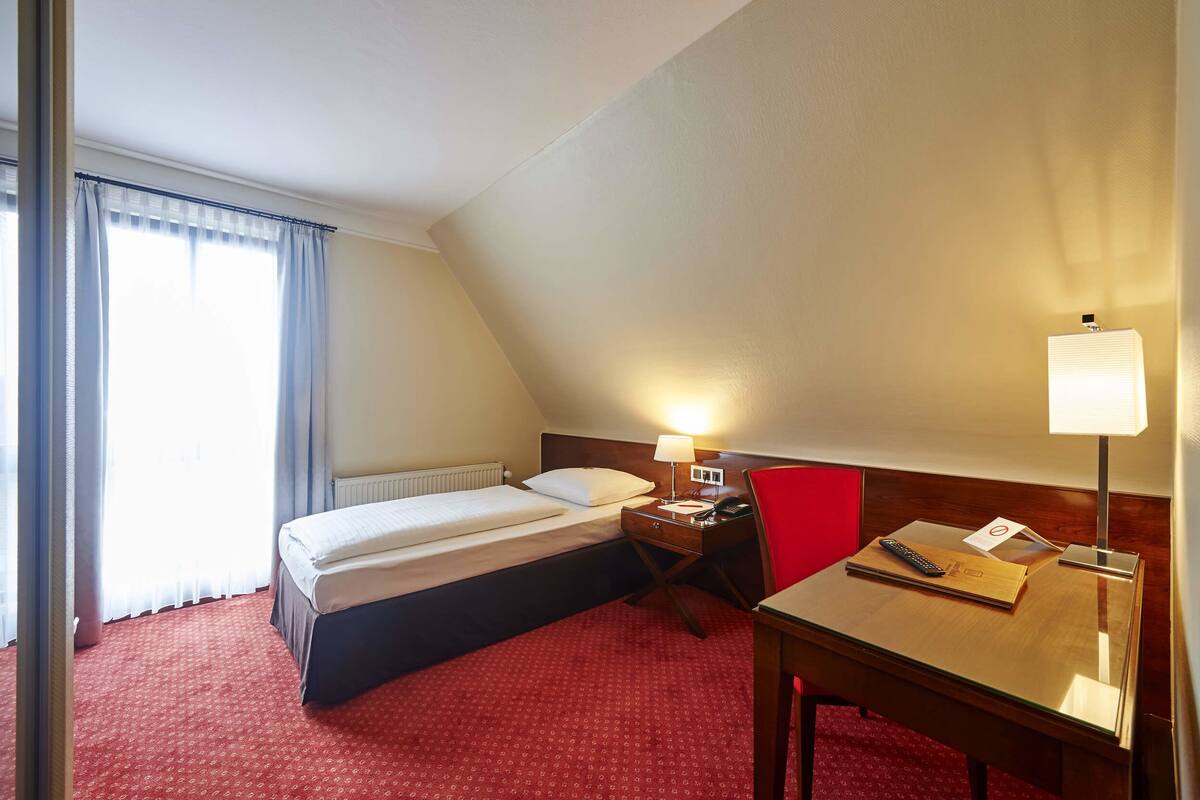 A hotel room with a bed, a desk, a chair, and a lamp.