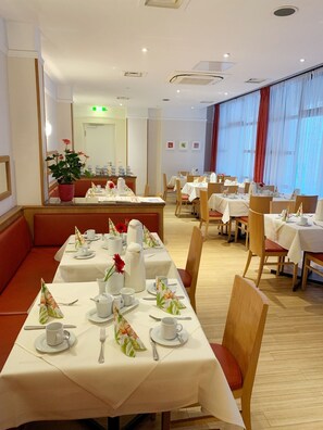 A restaurant with multiple dining tables set for guests, a counter with a cash register, and a large window with curtains. - Dürer Hotel Nürnberg (Nuremberg)