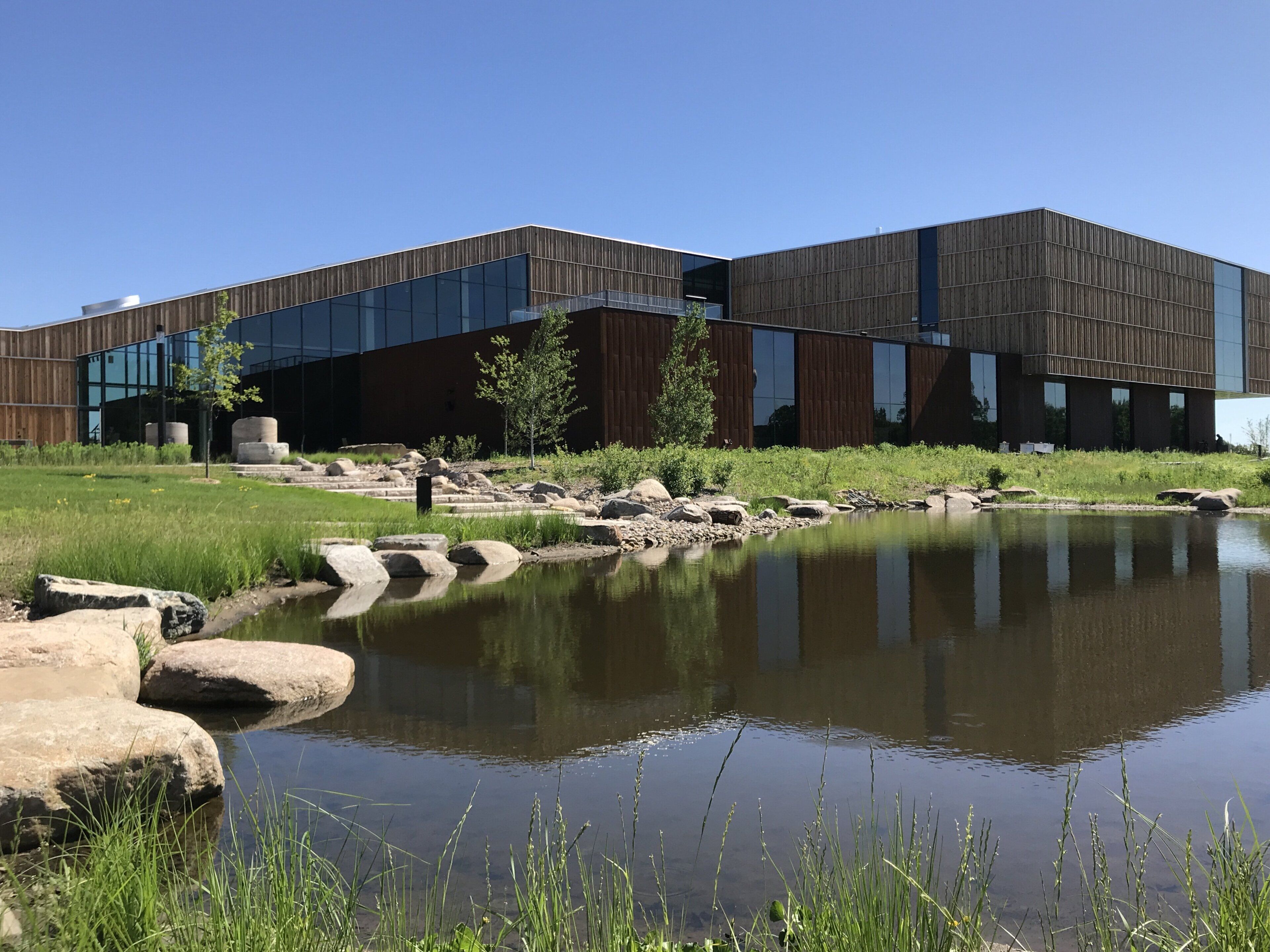 A modern building with a reflective glass facade, a landscaped area with a pond, and a clear blue sky.