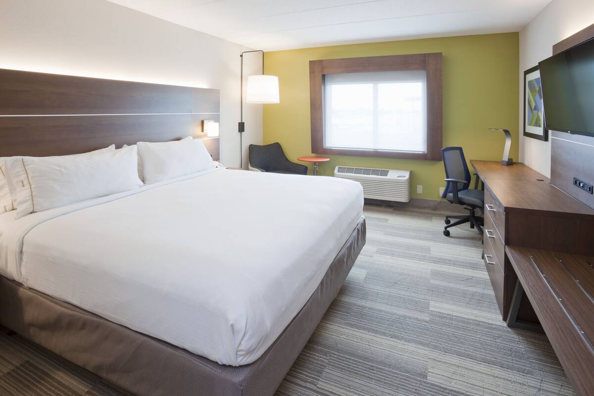 A hotel room with a large bed, a desk, a chair, a small table, an air conditioning unit, and a window with blinds.