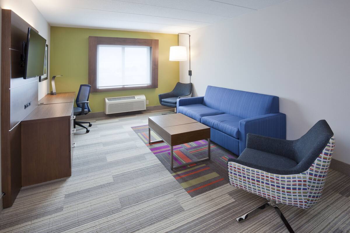 A hotel room with a wooden desk, a blue sofa, a patterned armchair, a television, and a window with a view.