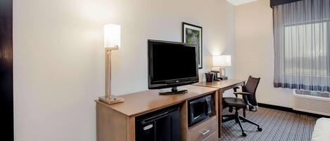 Habitación de hotel con un escritorio de madera, televisor de pantalla plana, un mini refrigerador, microondas, una silla, una lámpara y un ventanal con cortinas translúcidas.