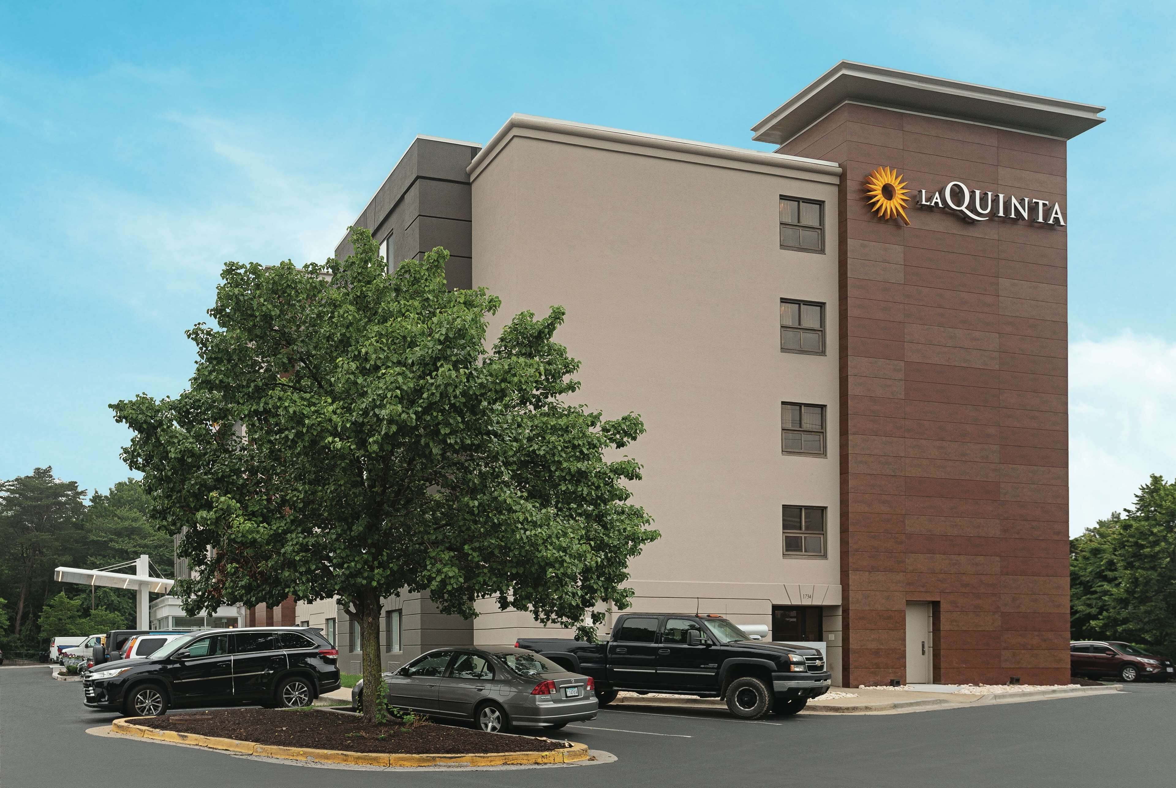A hotel named La Quinta with a modern design, featuring a parking lot with several vehicles and a large tree in front.