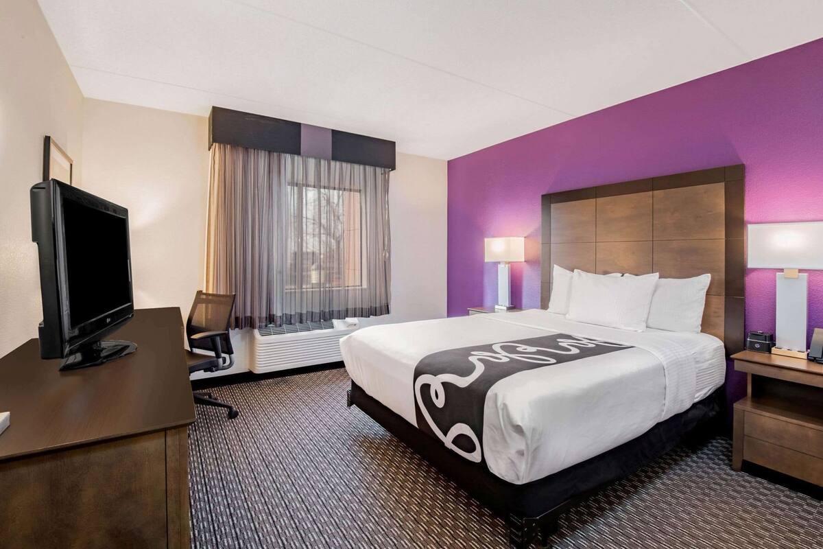 A hotel room with a bed, a desk with a TV, a window with curtains, and a nightstand with a lamp.