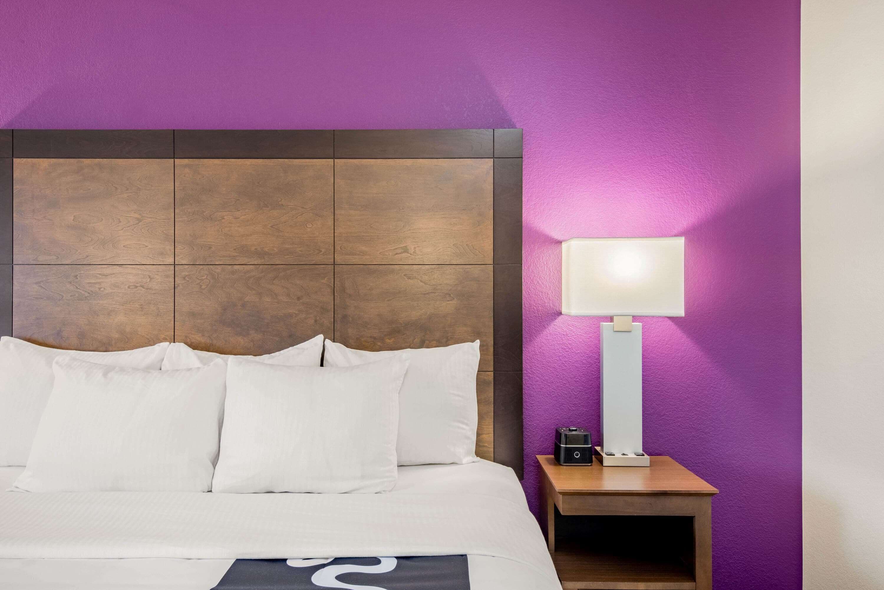 A hotel room with a wooden headboard, white bedding, a bedside table with a lamp, and a purple wall.