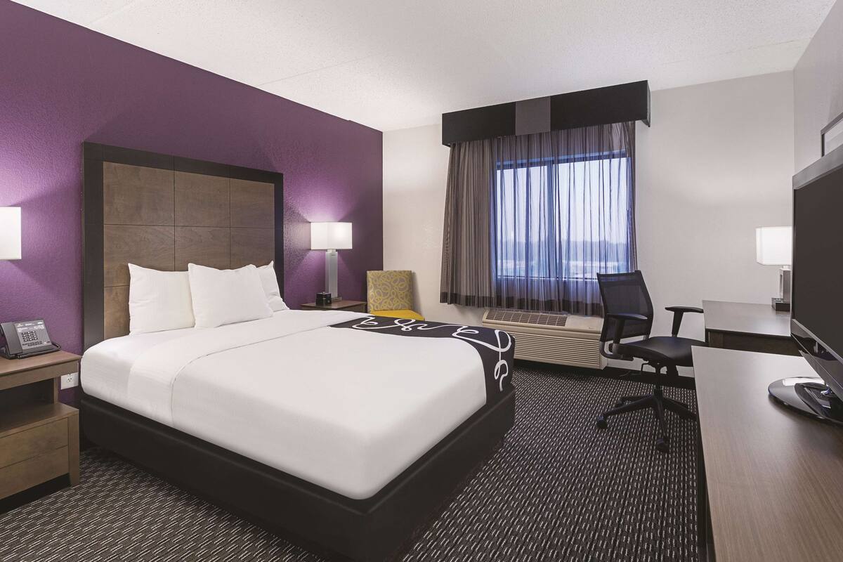 A hotel room with a bed, a desk, a chair, a lamp, and a window with curtains.