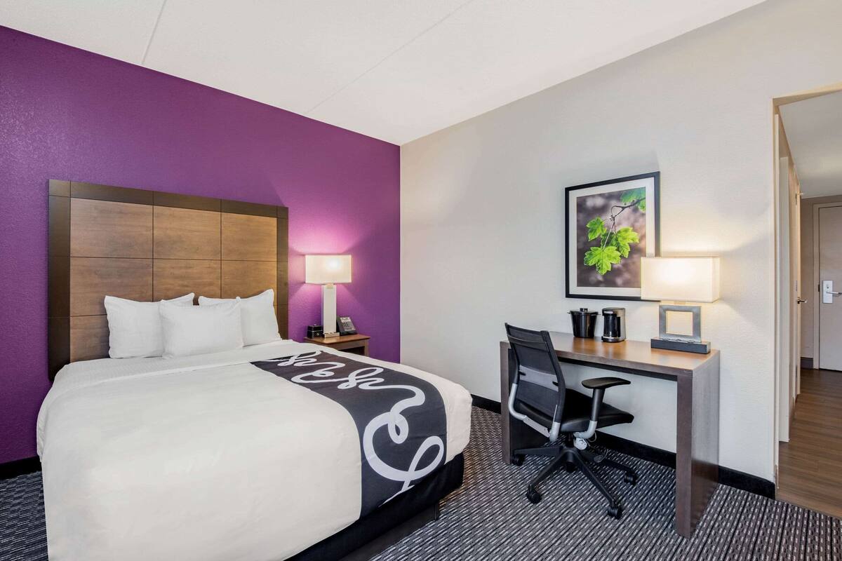 A hotel room with a bed, desk, chair, and a framed picture on the wall.