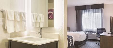 A hotel bathroom with a sink, mirror, and towels. The room has a bed, a desk, and a window with curtains.