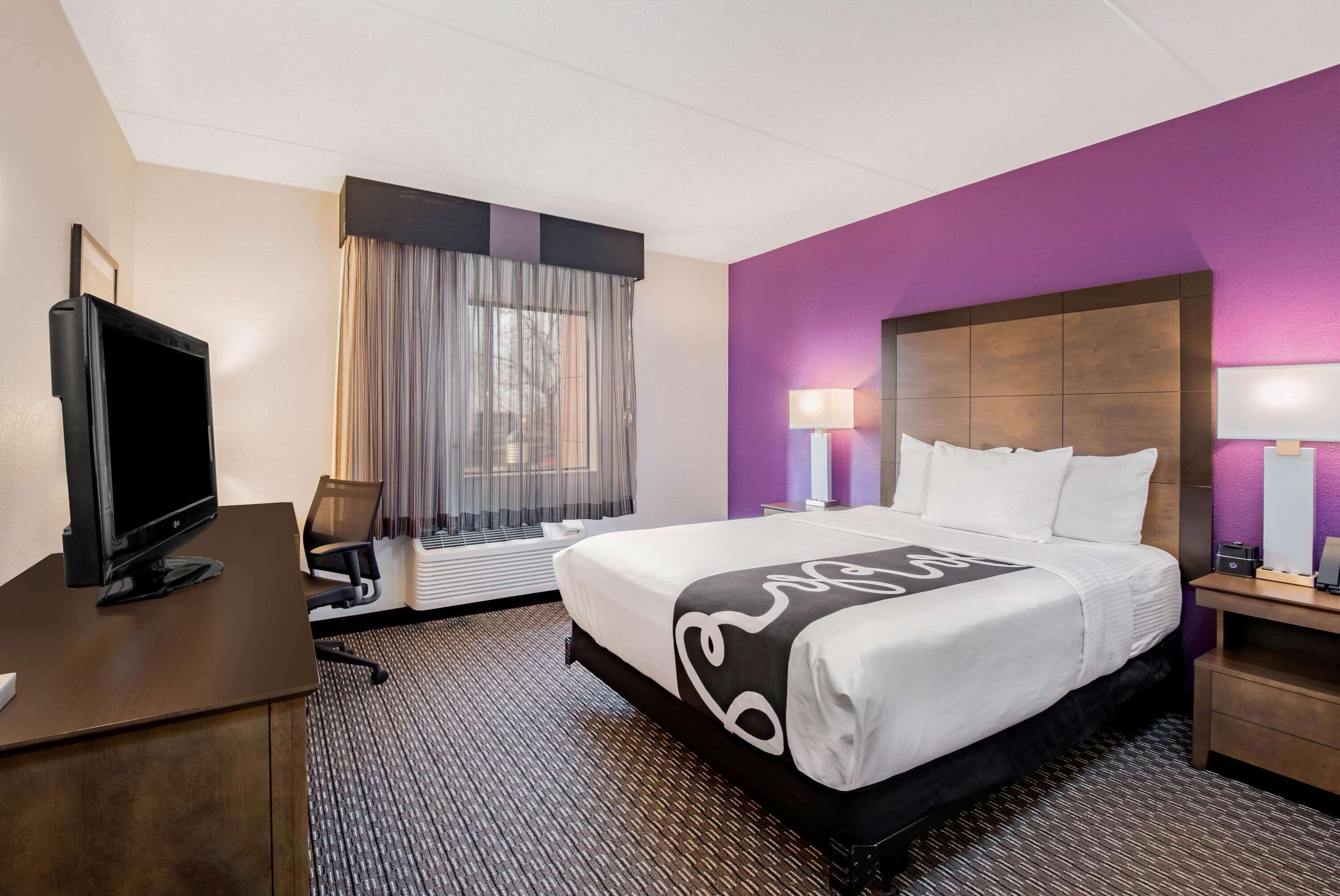 A hotel room with a large bed, a desk with a computer, a TV, and a window with curtains.