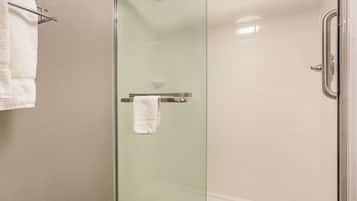 Ducha con cerramiento de vidrio, una toalla blanca en un perchero, paredes color beige y suelo de baldosas blancas.