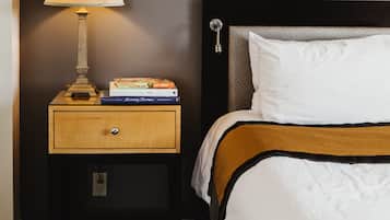 A neatly made bed with a white pillow and a brown and black striped blanket. A wooden nightstand with a lamp and a stack of books is beside the bed.
