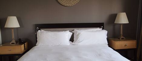 Una cama bien hecha con sábanas blancas, dos mesitas de noche con lámparas y un espejo decorativo en la pared.