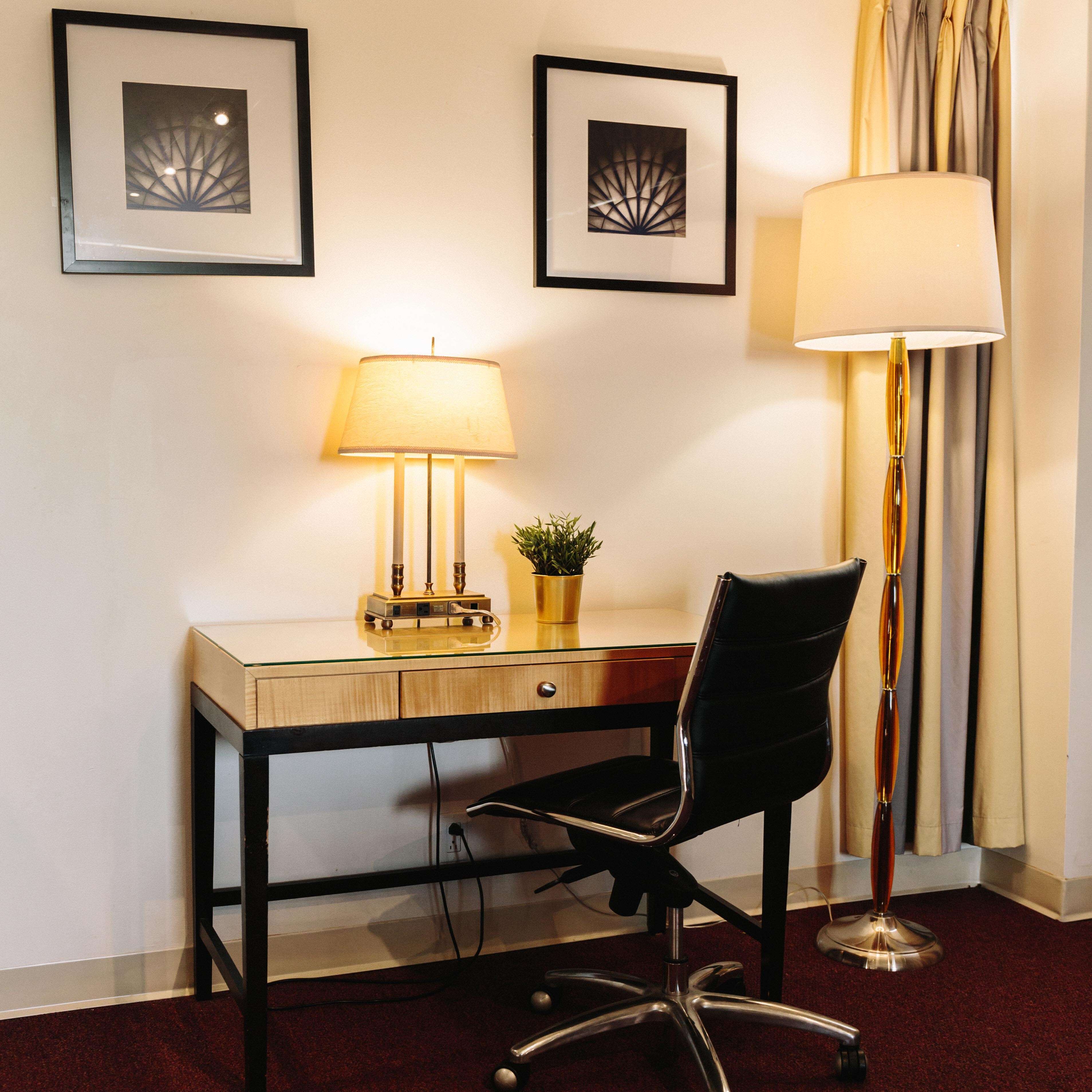 Ein Hotelzimmer mit einem Holztisch, einem schwarzen Bürostuhl, einer Stehlampe und zwei gerahmten Bildern an der Wand.