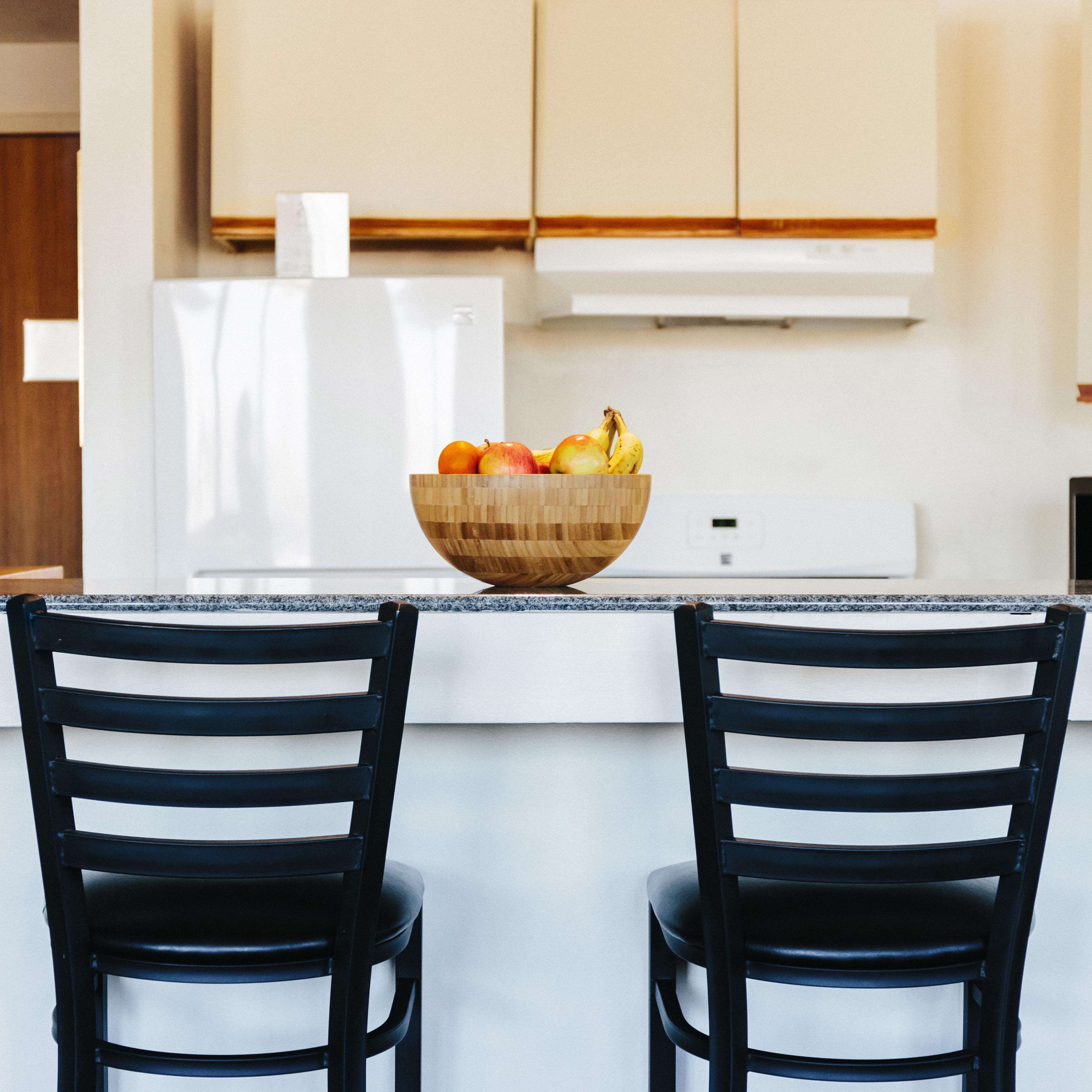 A modern kitchen with a wooden bowl of fruit on the island, two black chairs, and white cabinets.
